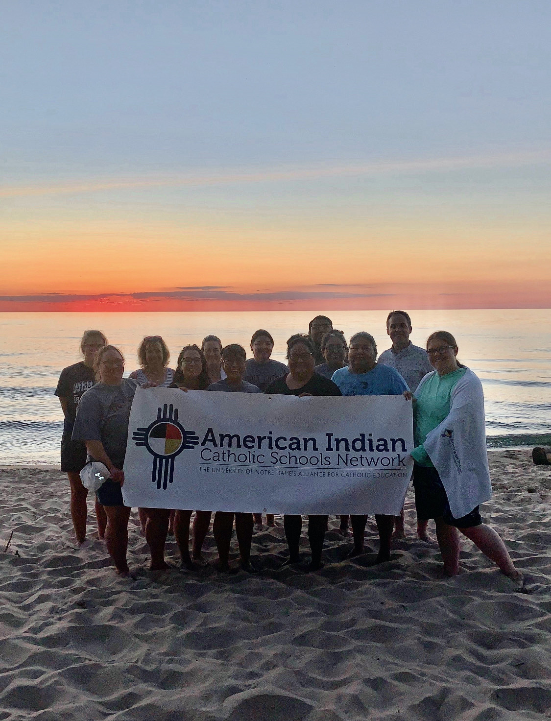 American Indian Catholic Schools Network group holding a sign with their program name at Lake Michigan on the beach.