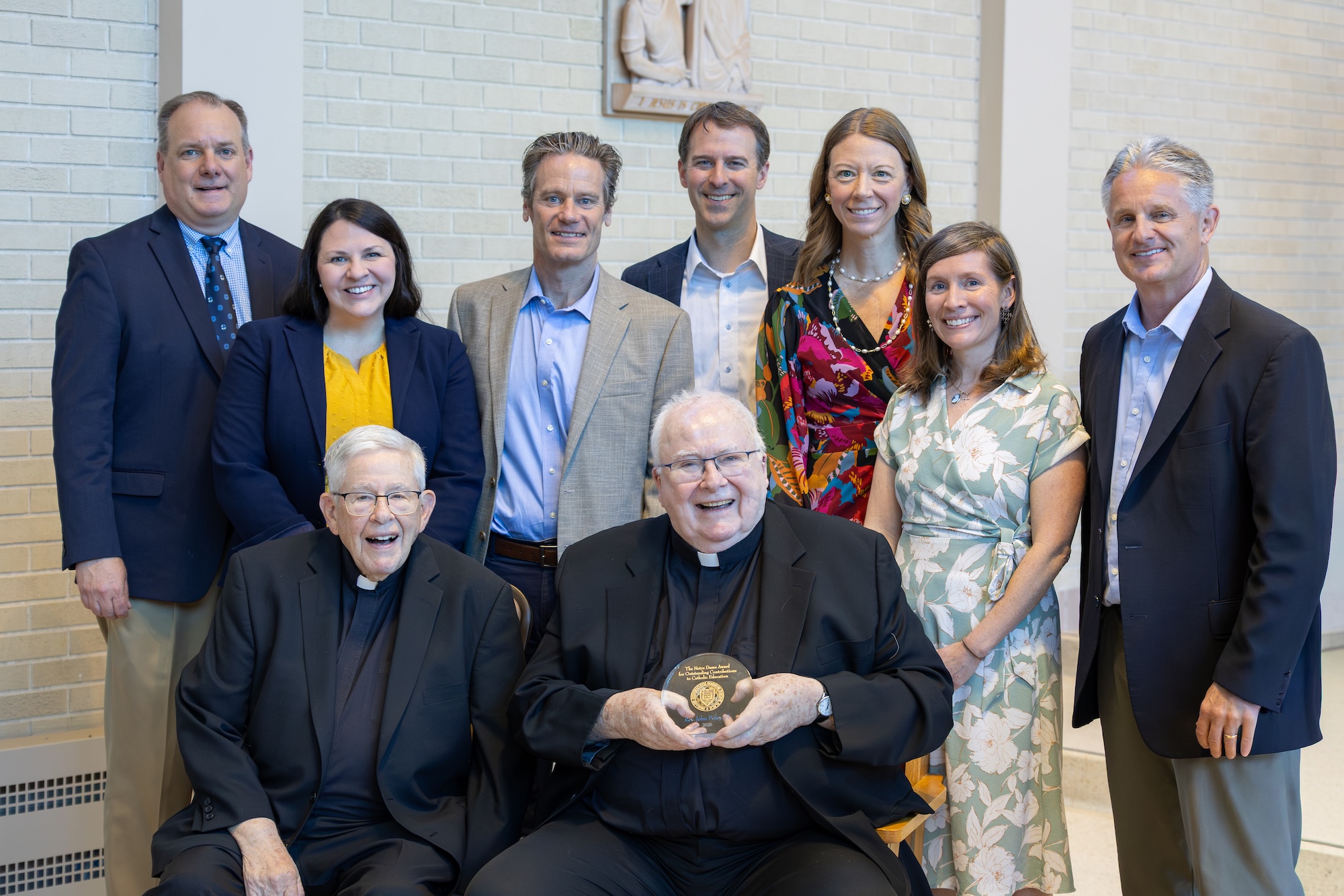 Priest Fr. John Foley seated at center holding award, surrounded by Notre Dame leadership