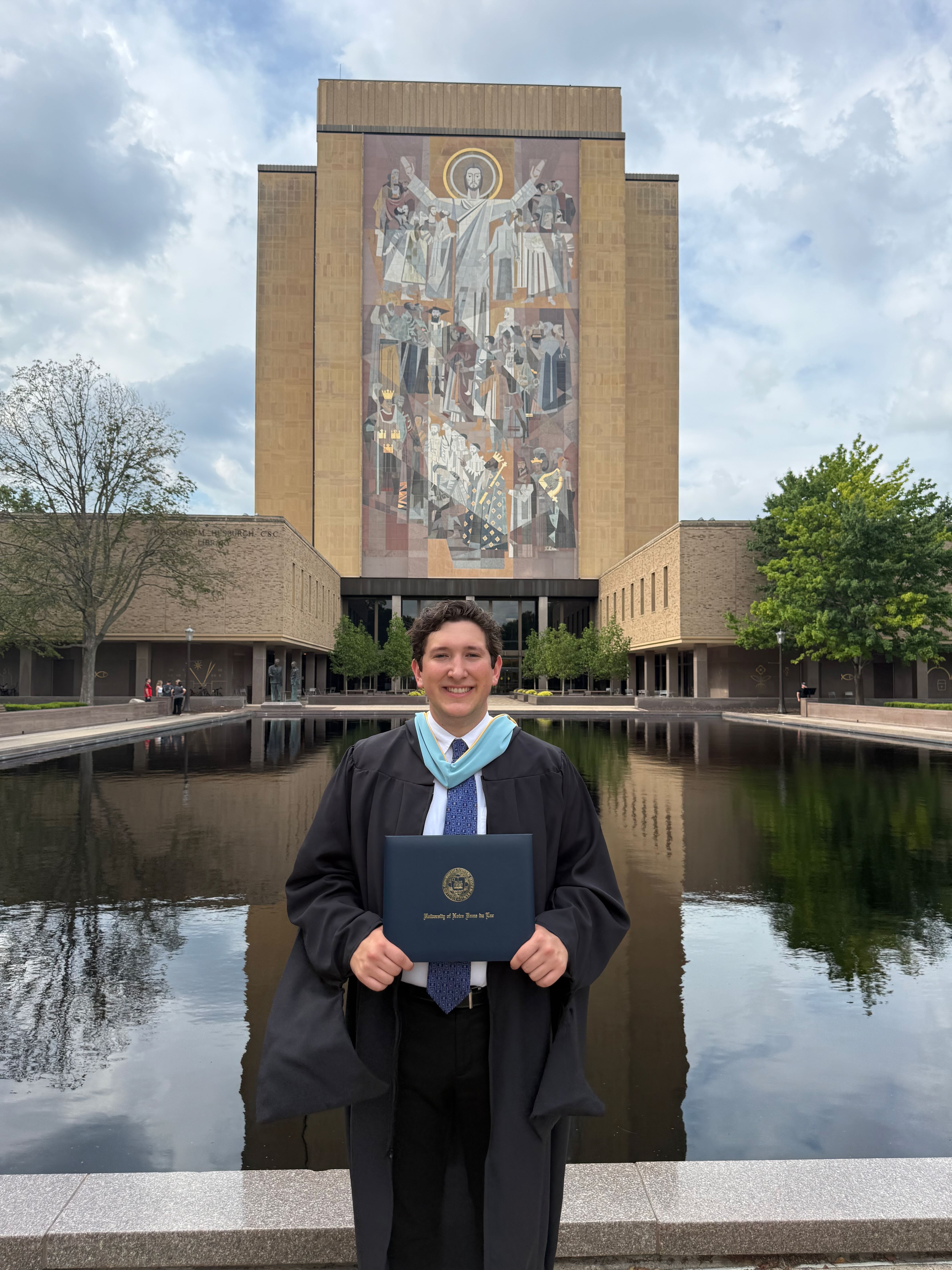 Image of Tom Betterly in his master's regalia in front of the Notre Dame Library and the Word of Life mural holding his diploma in his hands.