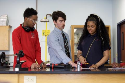 Clayton Glasgow (middle with blue button down and blue tie) talking to students on left (male in red sweater) and right (female in blue polo) or him. They are at a lab table looking at a worksheet on the table.