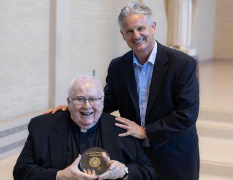Priest Fr. John Foley seated at center holding award, surrounded by Notre Dame leadership