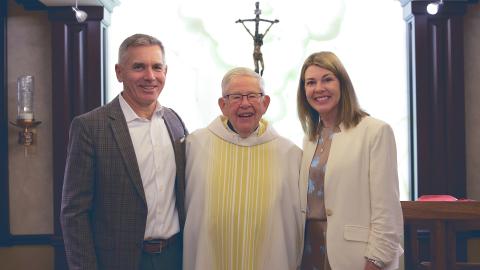 Elderly priest in white clerical robes stands between a smiling man in a dark grey suit and a smiling woman in a white jacket (right) 