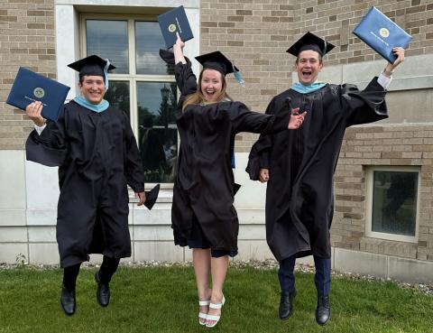 From left to right: Tom Betterly, Kate Jensen, and Sean Kelley in master's regalia holding their M.Ed. diploma in hand jumping up.