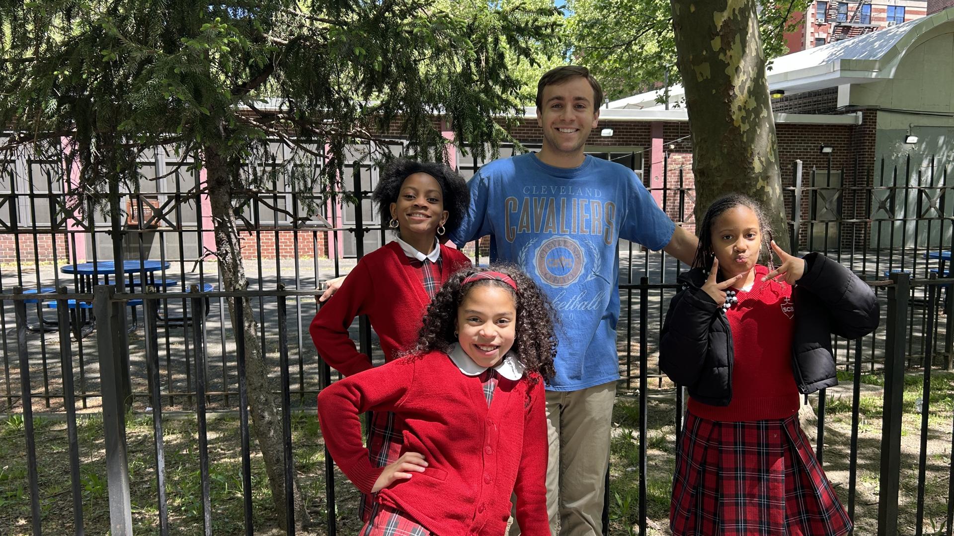 Image of Jack Hutter in blue tshirt posing with three of his students in red sweaters and red plaid skirts: two girls to his left and one to his right.