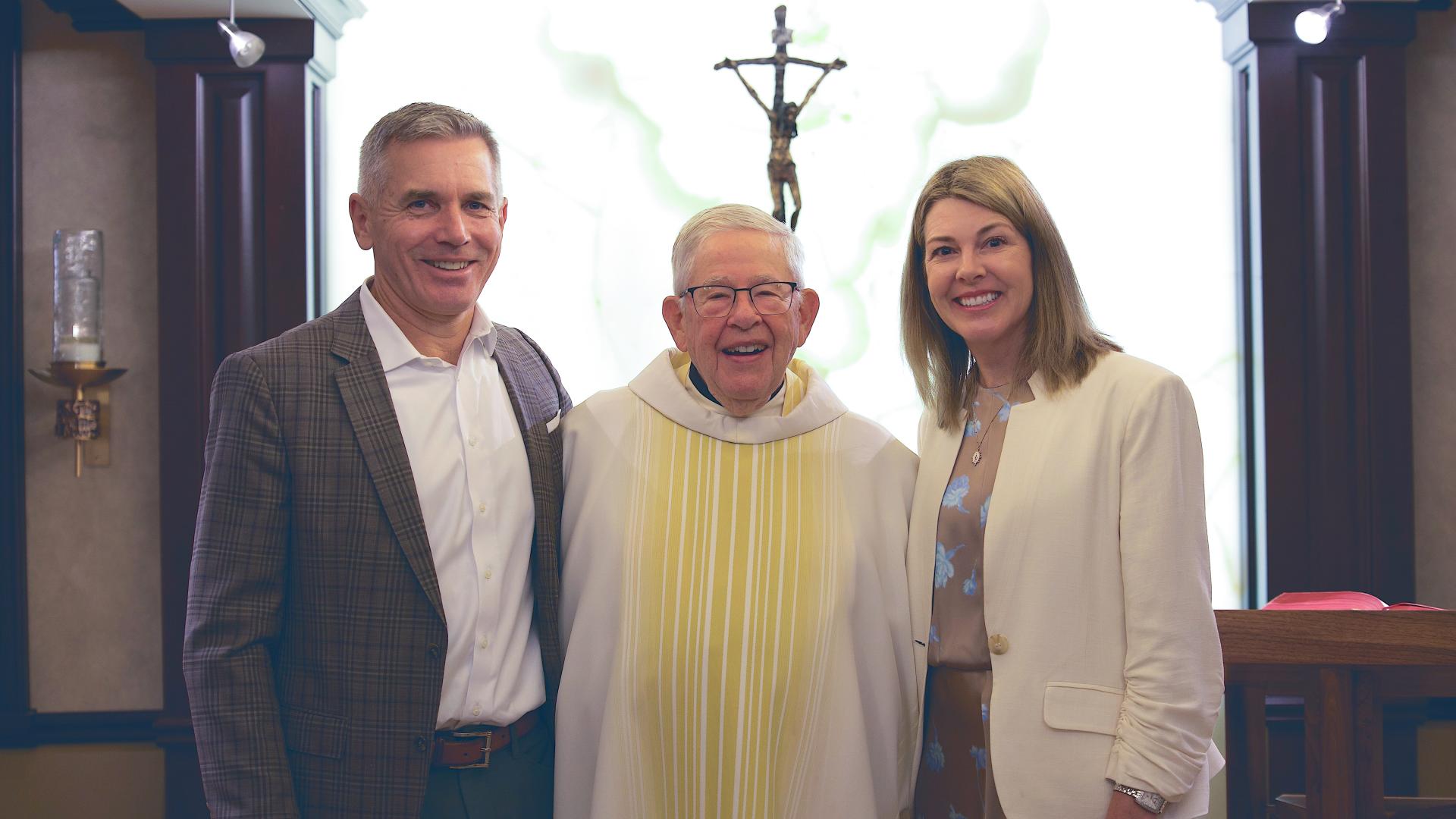 Elderly priest in white clerical robes stands between a smiling man in a dark grey suit and a smiling woman in a white jacket (right) 
