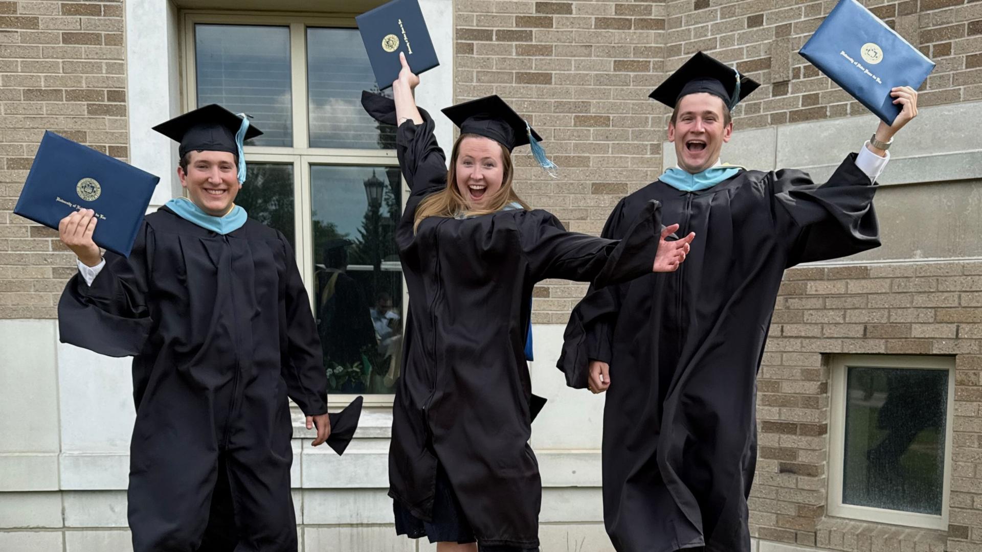 From left to right: Tom Betterly, Kate Jensen, and Sean Kelley in master's regalia holding their M.Ed. diploma in hand jumping up.